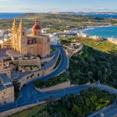 Malta, wyspa do kogo należy? Widok na kościół z czerwoną kopułą i kręte drogi na zielonym wzgórzu, z widokiem na morze i wybrzeże.