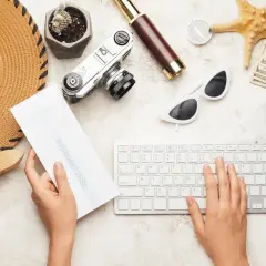 Ręce piszą na klawiaturze, trzymając kartę pokładową. Obok leżą kapelusz, aparat, okulary, pieniądze i kompas – idealne na podróż poślubną.