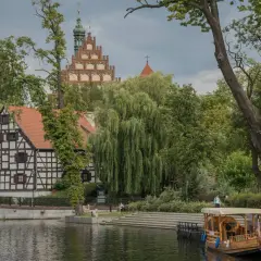 Nad wodą, wśród zieleni, malownicze kamieniczki i wieża tworzą magiczne miejsca kujawsko-pomorskie. Drewniana łódź czeka na podróżnych.