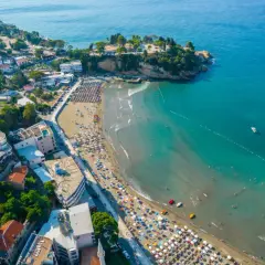 Majowa pogoda w Czarnogórze zachęca do plażowania. Widok z lotu ptaka na wybrzeże z turkusową wodą, piaszczystą plażą pełną leżaków i turystów.