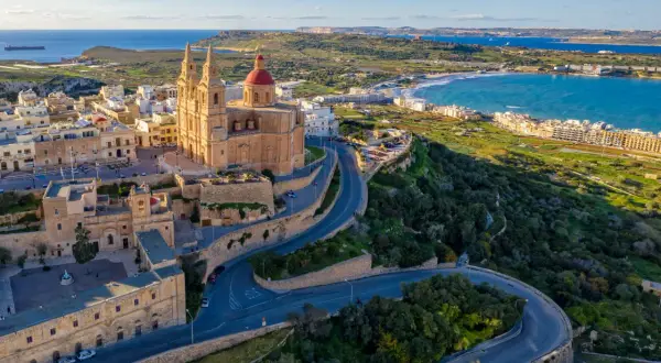 Malta, wyspa do kogo należy? Widok na kościół z czerwoną kopułą i kręte drogi na zielonym wzgórzu, z widokiem na morze i wybrzeże.
