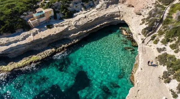 Malta plaże: turkusowa woda, skaliste wybrzeże i mały domek nad zatoczką. Dwie osoby spacerują ścieżką.