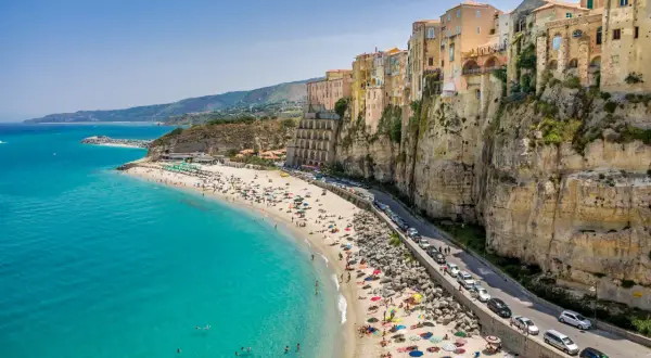 Tropea lotnisko: malownicza plaża z turkusową wodą, kolorowe budynki na klifie i droga z samochodami.