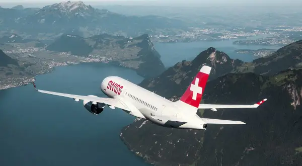 Samolot linii Helvetic Airways z charakterystycznym czerwonym ogonem i białym krzyżem leci nad malowniczym jeziorem i górami.