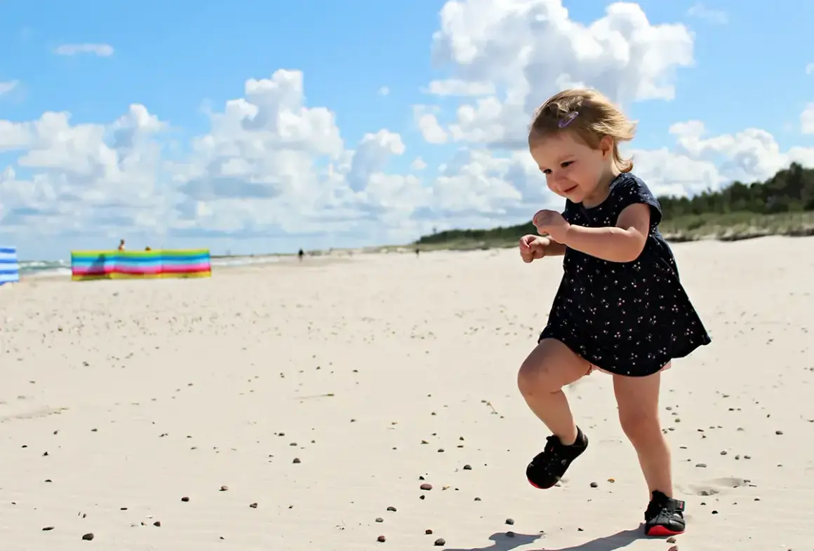 Radosne wakacje z dziećmi w Polsce. Mała dziewczynka biega po piaszczystej plaży, ciesząc się słońcem i chmurami na niebie.