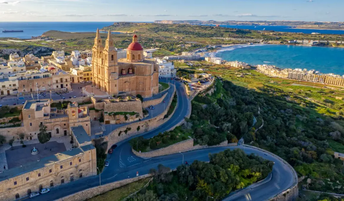 Malta, wyspa do kogo należy? Widok na kościół z czerwoną kopułą i kręte drogi na zielonym wzgórzu, z widokiem na morze i wybrzeże.