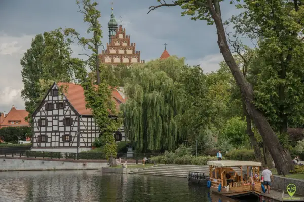 Nad wodą, wśród zieleni, malownicze kamieniczki i wieża tworzą magiczne miejsca kujawsko-pomorskie. Drewniana łódź czeka na podróżnych.