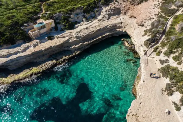 Malta plaże: turkusowa woda, skaliste wybrzeże i mały domek nad zatoczką. Dwie osoby spacerują ścieżką.