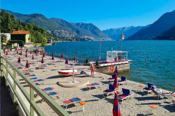 Plaże nad jeziorem Como, z rzędami leżaków i parasoli, czekają na turystów. W tle góry i błękitne niebo.
